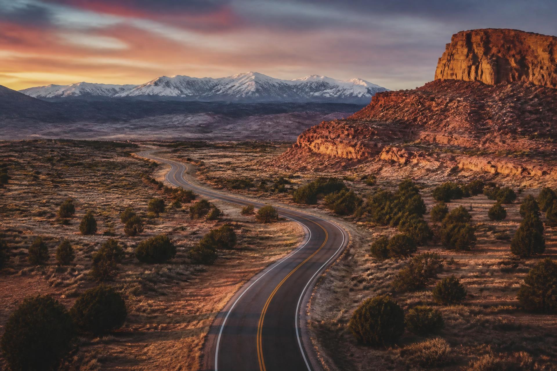 Empty highway winding through red rock mesas toward snow-capped mountains at sunset